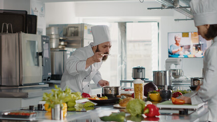 Professional chef testing soup taste in pot on stove, cooking delicious meal with sauce and vegetables. Confident man in uniform testing food recipe with utensils, preparing culinary menu.