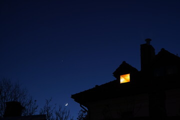 Silhouette of house roof with chimney and lit attic small window at night against clear sky with...