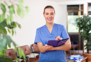 Cheerful smiling nurse woman standing in office with clipboard, writing medical history sheet