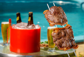 Brazilian barbecue picanha on skewers with blurred beers and pool in the background.