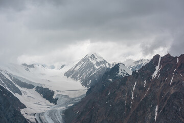 Fototapeta na wymiar Atmospheric landscape with sharp rocks and high snowy mountain top in rainy low clouds at overcast. Dramatic gloomy scenery with large snow mountains and glacier in gray cloudy sky at rainy weather.