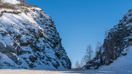 The road runs between steep cliffs with snow-covered slopes. Ahead - clear blue sky, bare trees. Altai