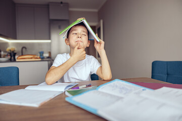 Cute curious Caucasian small schoolboy holding textbook over head with guessing face.