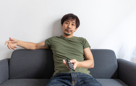 Bored Adult Asian Man Watching Television Sitting On Couch At Home.