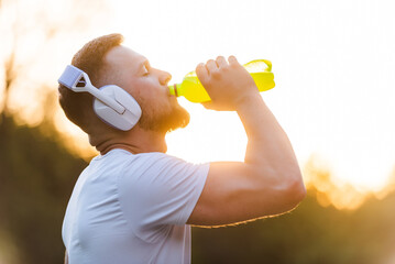 Young sports man drinking energy water from a bottle while resting after run or workout on sunlight...