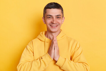 Indoor shot of positive man wearing casual style hoodie, posing isolated over yellow background, standing in yoga pose and try to relaxing, keeps palms together.