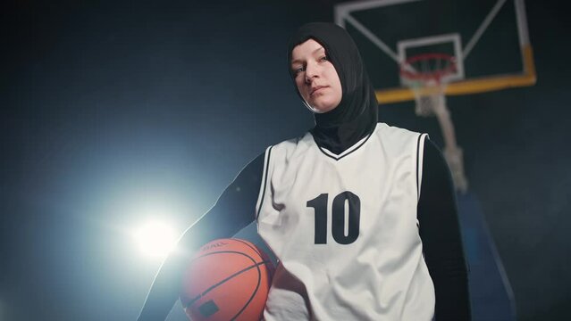 Slow motion, portrait of a young arab woman basketball player, holding a ball in her hands and looking at the camera, women's basketball championship, serious look.