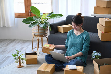 Smiling young woman using laptop and preparing parcel boxes of product for delivery to customers....