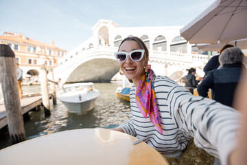 Fototapeta na wymiar Young woman making selfie on phone while sitting at outdoor cafe near Grand Canal and Rialto bridge in Venice. Caucasian traveler resting at italian cafe. Idea of a happy vacation in Italy