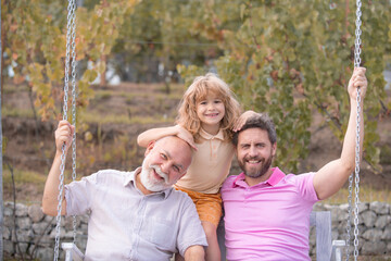 Three generations of men together, portrait of smiling son, father and grandfather swinging on the swing, having fun in the park outdoor.