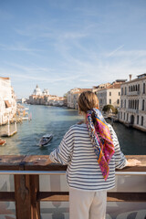 Fototapeta na wymiar Young woman enjoying beautiful view on Grand Canal from Academy bridge in Venice. Idea of spending summer time and travel in Italy. Caucasian female wearing striped vest and colorful shawl