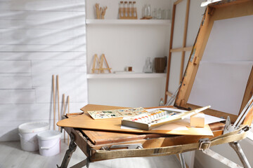 Foldable wooden easel with empty canvas and supplies in studio. Artist's workplace