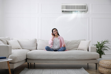 Young woman resting under air conditioner on white wall at home