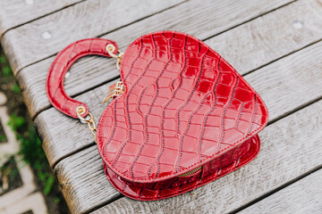 A red women's handbag in the form of a heart lies on a bench in a city park