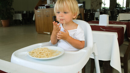 Baby boy eats spaghetti sitting in a baby chair in restaurant