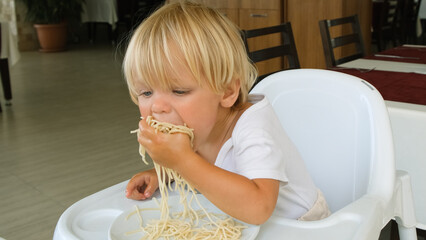Baby boy eats spaghetti sitting in a baby chair in restaurant