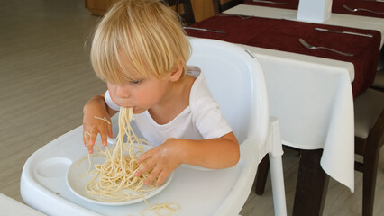 Baby boy eats spaghetti sitting in a baby chair in restaurant