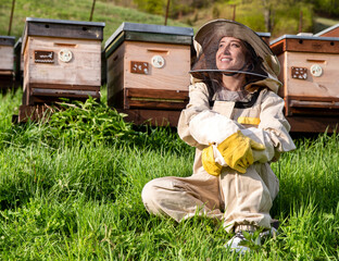 Girl beekeeper in protective white suit posing in front of wooden beehives. Young apiarist