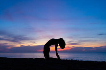 Silhouette of a beautiful woman practicing yoga asana on the beach with sunrise on twilight blue vibrant sky and calm sea in background. Camel pose, Ustrasana, June 21, International Yoga Day, Concept