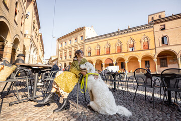 Stylish woman sitting with her white dog at outdoor cafe in the old town of Bologna city. Italian...