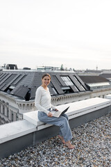 Fototapeta na wymiar cheerful woman in jeans sitting on rooftop with laptop and smiling at camera