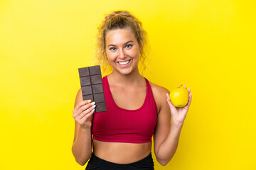 Girl with curly hair isolated on yellow background taking a chocolate tablet in one hand and an...