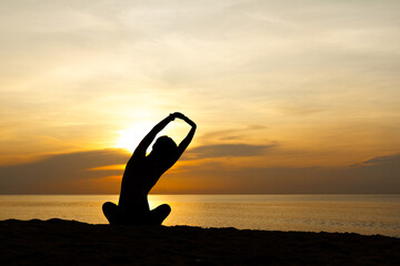 Silhouette of beautiful woman sitting cross legged and stretching her arms up to sky, sunrise on...