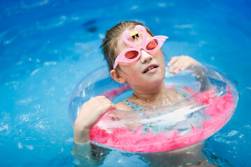 Obraz na płótnie Canvas european preteen girl in flamingo sunglasses and a swimsuit swims on circle in pool in backyard. Funny child in pink glasses with shells and with an inflatable ring with pink feathers