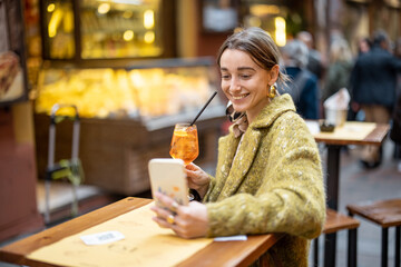 Happy woman having video call or taking selfie photo on phone while sitting with cocktail at...