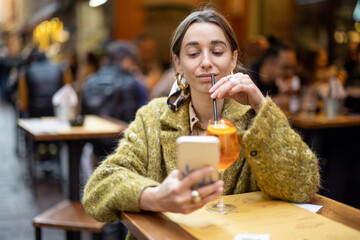 Young woman using phone while sitting with spritz aperol drink at bar outdoors. Concept of Italian...