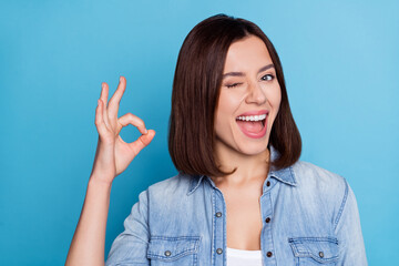 Photo of positive funky girl eye wink fingers show okey symbol toothy smile isolated on blue color...