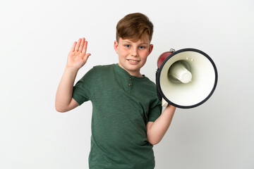 Little redhead boy isolated on white background holding a megaphone and saluting with hand with happy expression