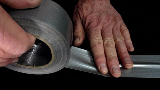 Closeup ground level shot of a man’s hands pulling duct tape from a roll and pressing to stick it over a white power cable, lying in a straight line across a black floor. 