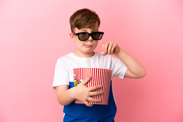 Little redhead boy isolated on pink background with 3d glasses and holding a big bucket of popcorns