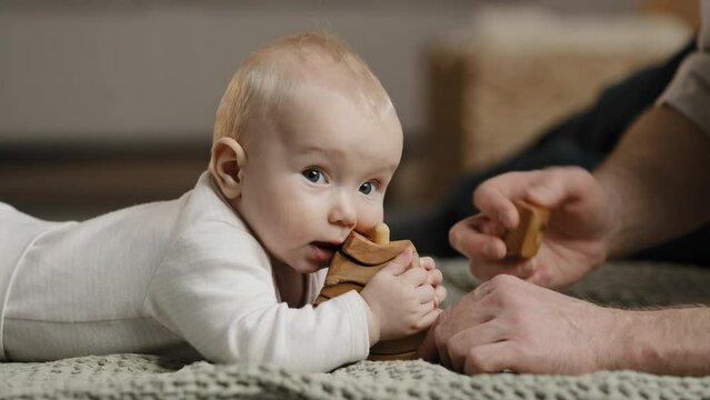 Caucasian family unrecognizable father daddy play with little daughter son infant newborn toddler lying at home on floor with toys baby bites wooden constructor teething concept, man stroking head