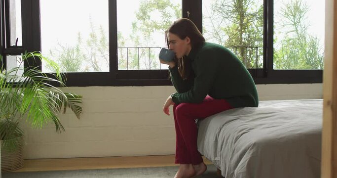 Caucasian non-binary transgender woman sitting on bed and drinking coffee