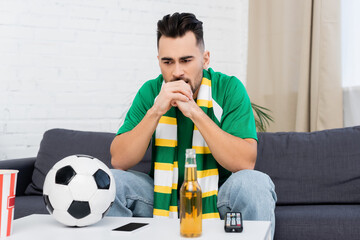 frustrated sports fan sitting near tv remote controller, soccer ball and beer.