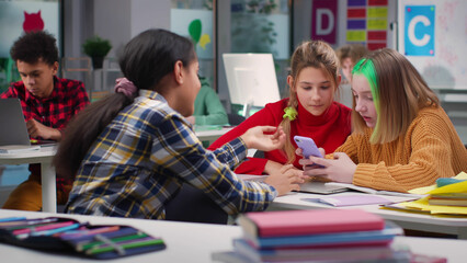 Fototapeta na wymiar Diverse teen kids at school discuss content on cellphone sitting in classroom
