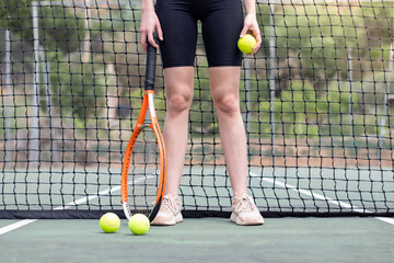 Front view of unrecognizable tennis player woman legs and hands in front of a net holding a ball and a racket on a court