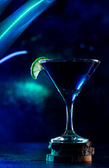 a cocktail glass with a drink inside stands on a stand with a slice of lime on the edge. neon trail behind the shot glass