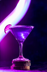 a cocktail glass with a drink inside stands on a stand with a slice of lime on the edge. neon trail behind the shot glass