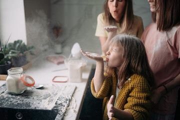 A lesbian family with a daughter are standing in the kitchen and blowing flour off their hands