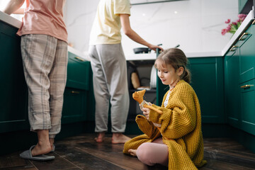 A lesbian family is doing household chores in the kitchen, and their daughter in a yellow sweater is sitting on the floor and playing with a toy