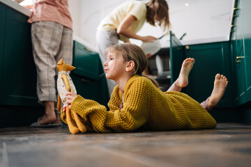 A lesbian family is doing household chores in the kitchen, and their daughter in a yellow sweater is lying on the floor and playing with a toy