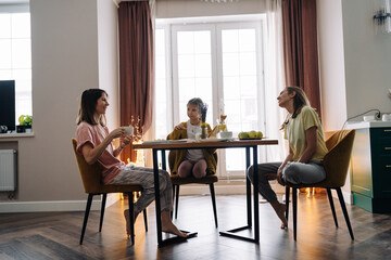 Fototapeta na wymiar Lesbian family with their daughter sitting at the kitchen table, having breakfast and smiling