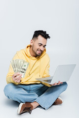 happy bookmaker sitting with dollar banknotes and laptop on grey.