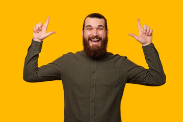 Portrait of a bearded man pointing up with both hands at the copy space. Studio shot over yellow background.