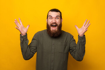Young bearded man is shocked and amazed while gesturing at the camera. Studio shot over yellow background.