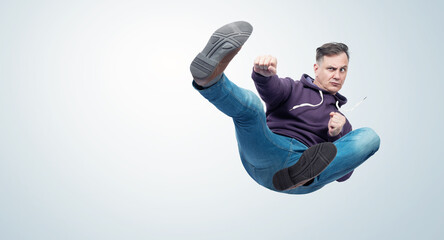Man in dark violet hooded sweatshirt and jeans in karate fight jump, on light blue background.