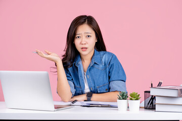 Young asian woman is confused mistaken disappointed she wearing casual shirt sit work at white office desk with laptop and spreading hands isolated on pastel pink background studio